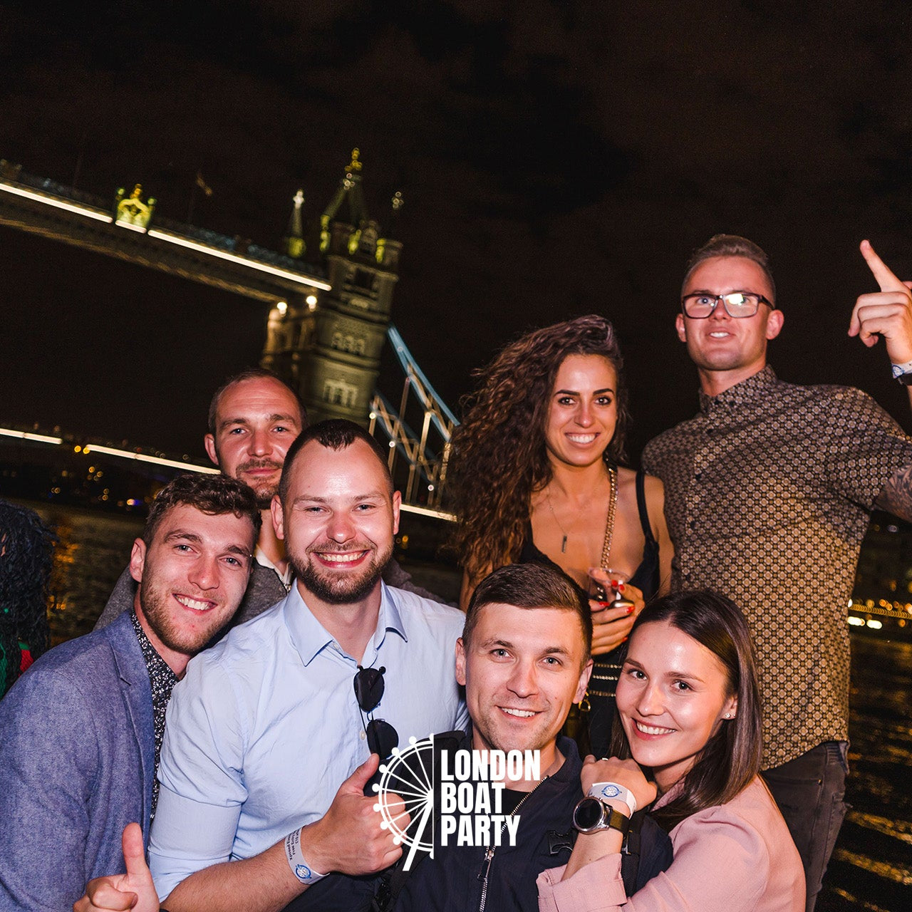 Group of people posing for a photo at a London boat party with Tower Bridge in the background.