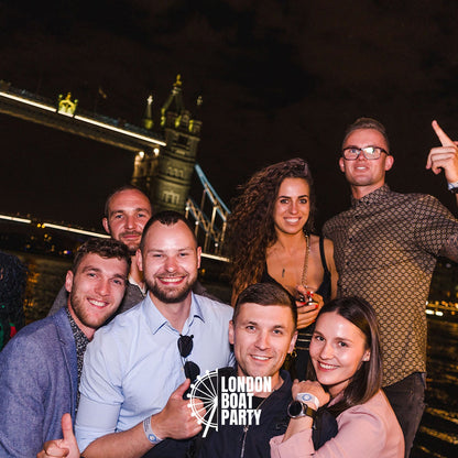 Group of people posing for a photo at a London boat party with Tower Bridge in the background.
